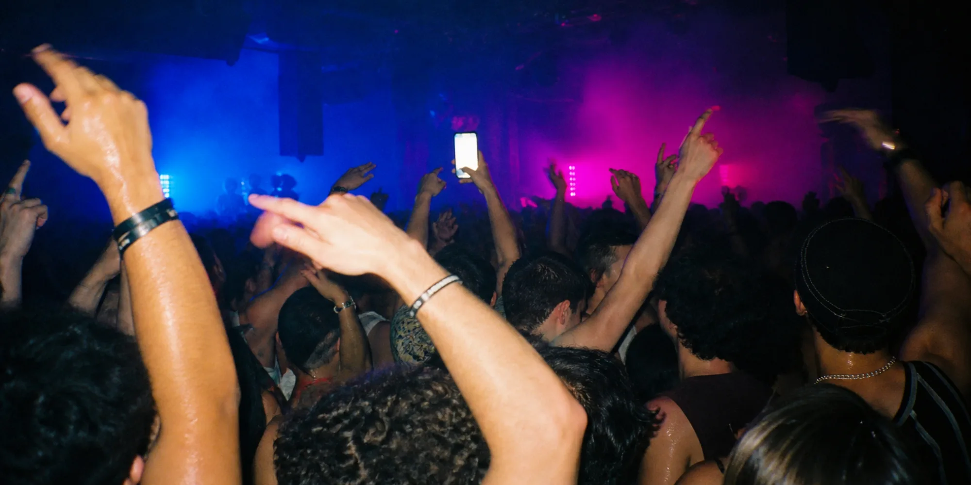 A packed venue at night, arms raised, one phone held above the crowd with the screen glowing in blue and magenta stage haze.