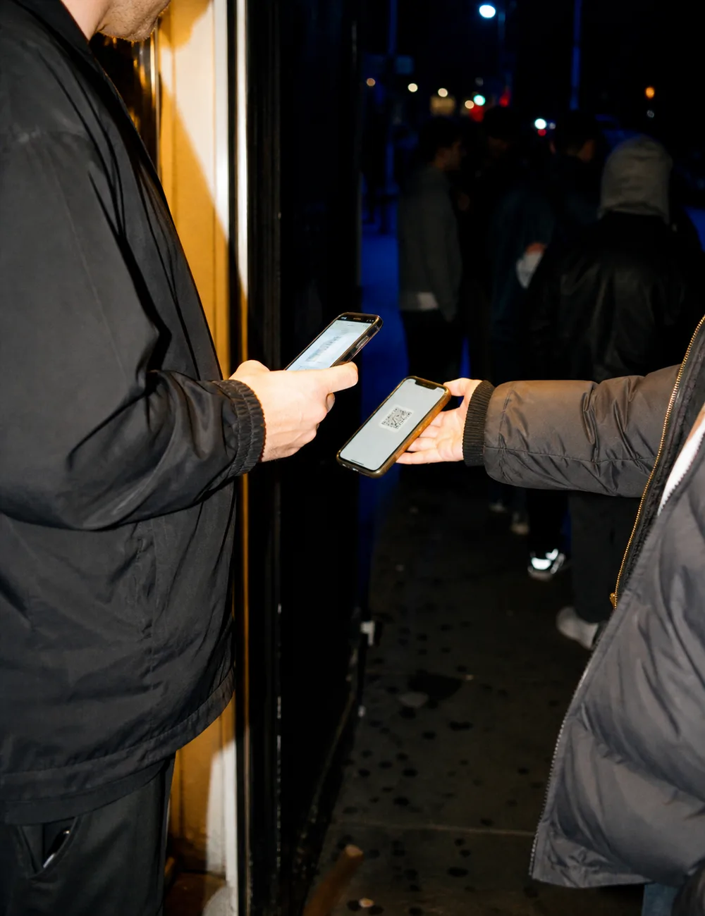 A door host at a venue entrance at night scanning the QR ticket on a guest phone, with a queue waiting under warm doorway light.