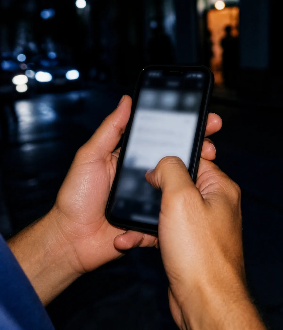 Close crop of two hands holding a phone on a street at night, the screen glowing, a thumb mid-tap.